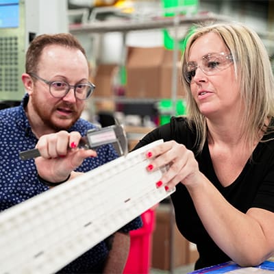 An AJAC machinist apprentice measures a part with a caliper as Evan Love, Workforce Development Specialist looks on.